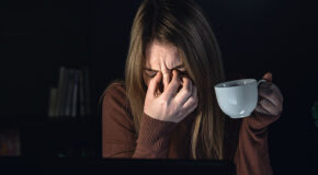 En trøtt kvinne holder en kopp kaffe mens hun kniper seg i øynene