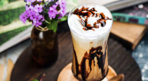 Et høyt glass med iskaffe med nutella står på et serveringsfat ved siden av en blomstervase