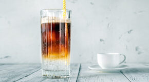 Et glass med mocktailen coffee sundown står på en benk med en hvit kaffekopp i bakgrunnen