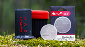 Et AeroPress-metallfilter ligger på mose ved siden av en AeroPress og en AeroPress Go
