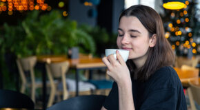En ung kvinne med lukkede øyne sitter på kafé og holder en kopp kaffe opp mot nesa