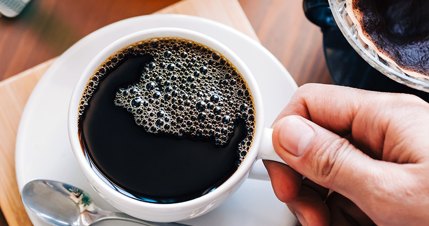 En hånd løfter en kopp med svart kaffe opp fra en skål