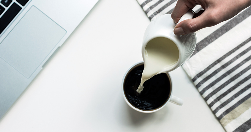 En person heller melk fra en liten mugge oppi en kopp med kaffe