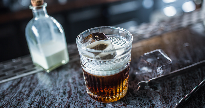 En white russian-cocktail står på en bardisk ved siden av en mugge med melk