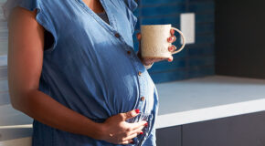 En gravid kvinne holder en kopp kaffe i én hånd og har den andre hånden på magen