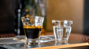 Et glass med espresso står ved siden av et lite vannglass