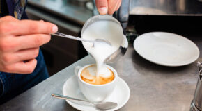 En person bruker en skje til å dytte melkeskum fra en pitcher over i en kaffedrikk