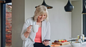 En eldre kvinne drikker kaffe og smiler mens hun ser på mobilen sin