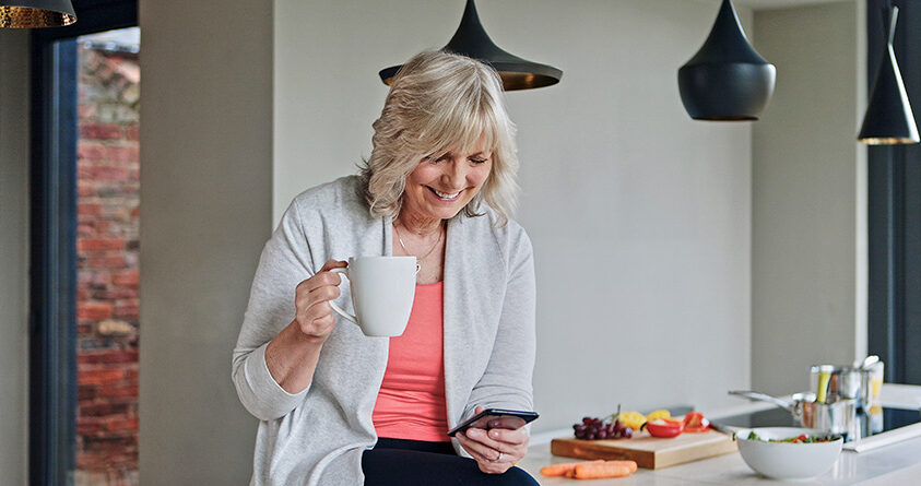 En eldre kvinne drikker kaffe og smiler mens hun ser på mobilen sin