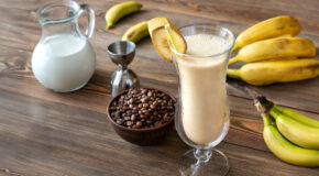 Et glass med kaffesmoothie står på en kjøkkenbenk ved siden av kaffebønner og et par bananklaser