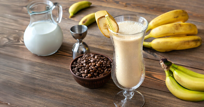Et glass med kaffesmoothie står på en kjøkkenbenk ved siden av kaffebønner og et par bananklaser