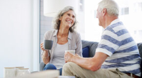 Et eldre ektepar ser smilende på hverandre mens de holder en kopp kaffe