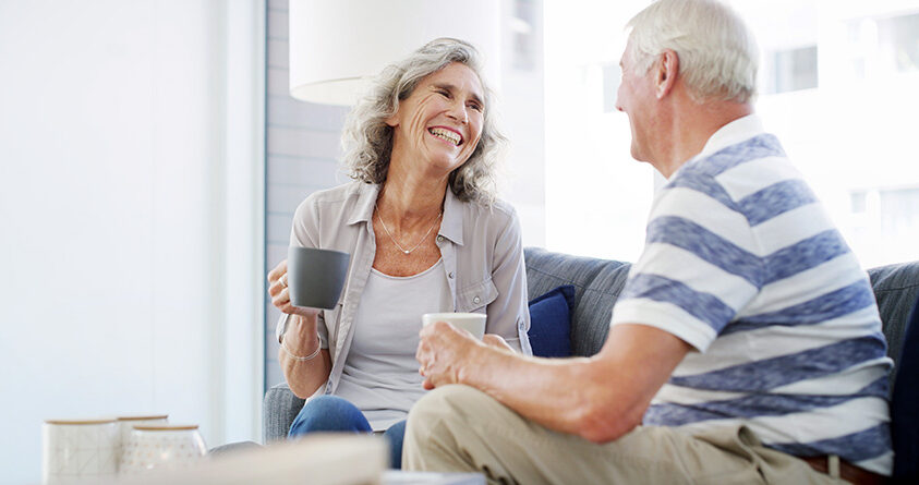 Et eldre ektepar ser smilende på hverandre mens de holder en kopp kaffe