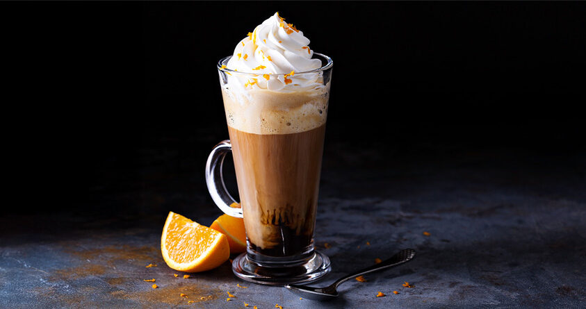 Et glass med mocca står på en mørk benk ved siden av en oppkuttet appelsin