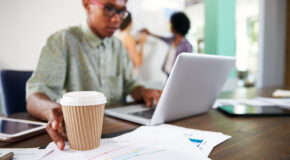 En mann strekker seg etter en kopp med kaffe mens han jobber på en laptop