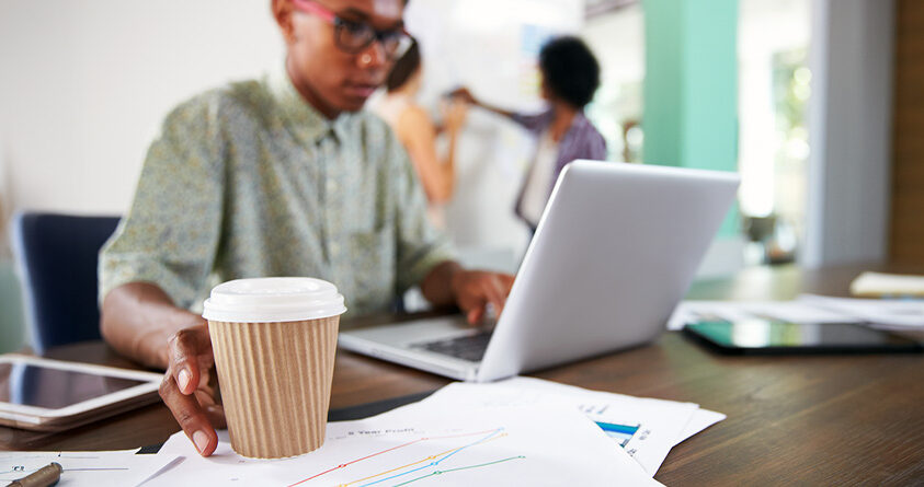 En mann strekker seg etter en kopp med kaffe mens han jobber på en laptop