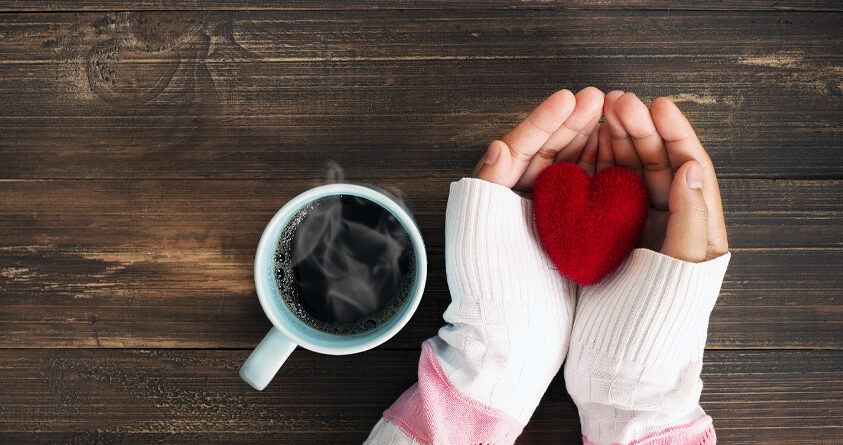 En person holder et rødt stoffhjerte mellom hendene, ved siden av en kopp med svart kaffe