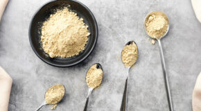 En skål og fire teskjeer med maca-pulver ligger på et grått underlag