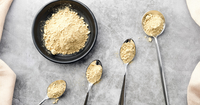 En skål og fire teskjeer med maca-pulver ligger på et grått underlag
