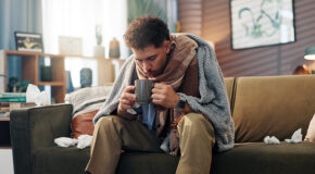 En forkjølet mann sitter pakket inn i pledd på en sofa og holder en kopp kaffe