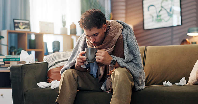 En forkjølet mann sitter pakket inn i pledd på en sofa og holder en kopp kaffe