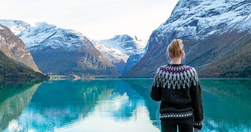 En kvinne med ullgenser står foran et vann, omringet av norske fjell