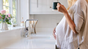En gravid kvinne står ved kjøkkenbenken og holder en kopp med kaffe