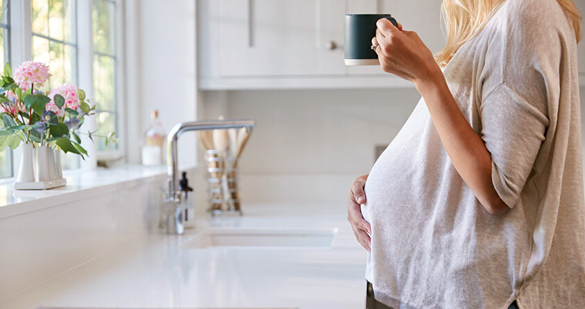 En gravid kvinne står ved kjøkkenbenken og holder en kopp med kaffe