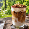 Et glass med tiramisu-islatte står på et bord ute i en grønn hage